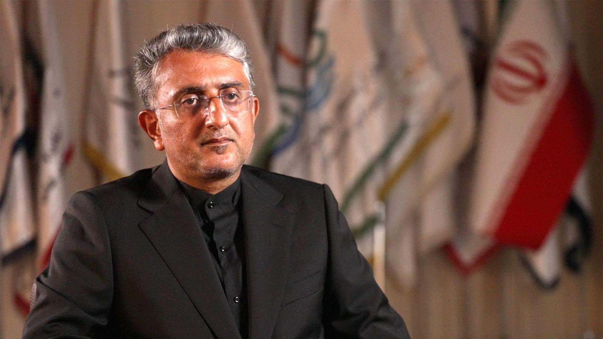 A man wearing a black shirt and jacket looks at the camera. He is sitting down with flags behind him, one of which is the Iranian flag. 