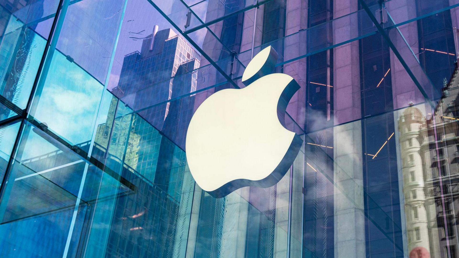 The Apple logo on the side of a building. It is a cartoon of an apple with a bite taken out of it. The building is reflecting lots of colours from the surrounding area like purple, blue and green.