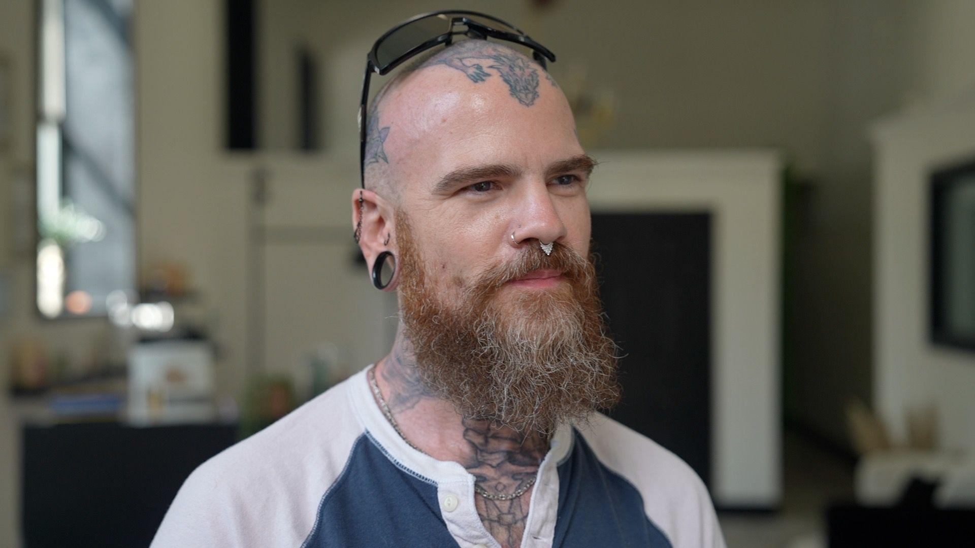 Bald and bearded Adam Teague, 44, wears sunglasses atop his head, which is tatooed, along with his neck, and wears two nose piercings