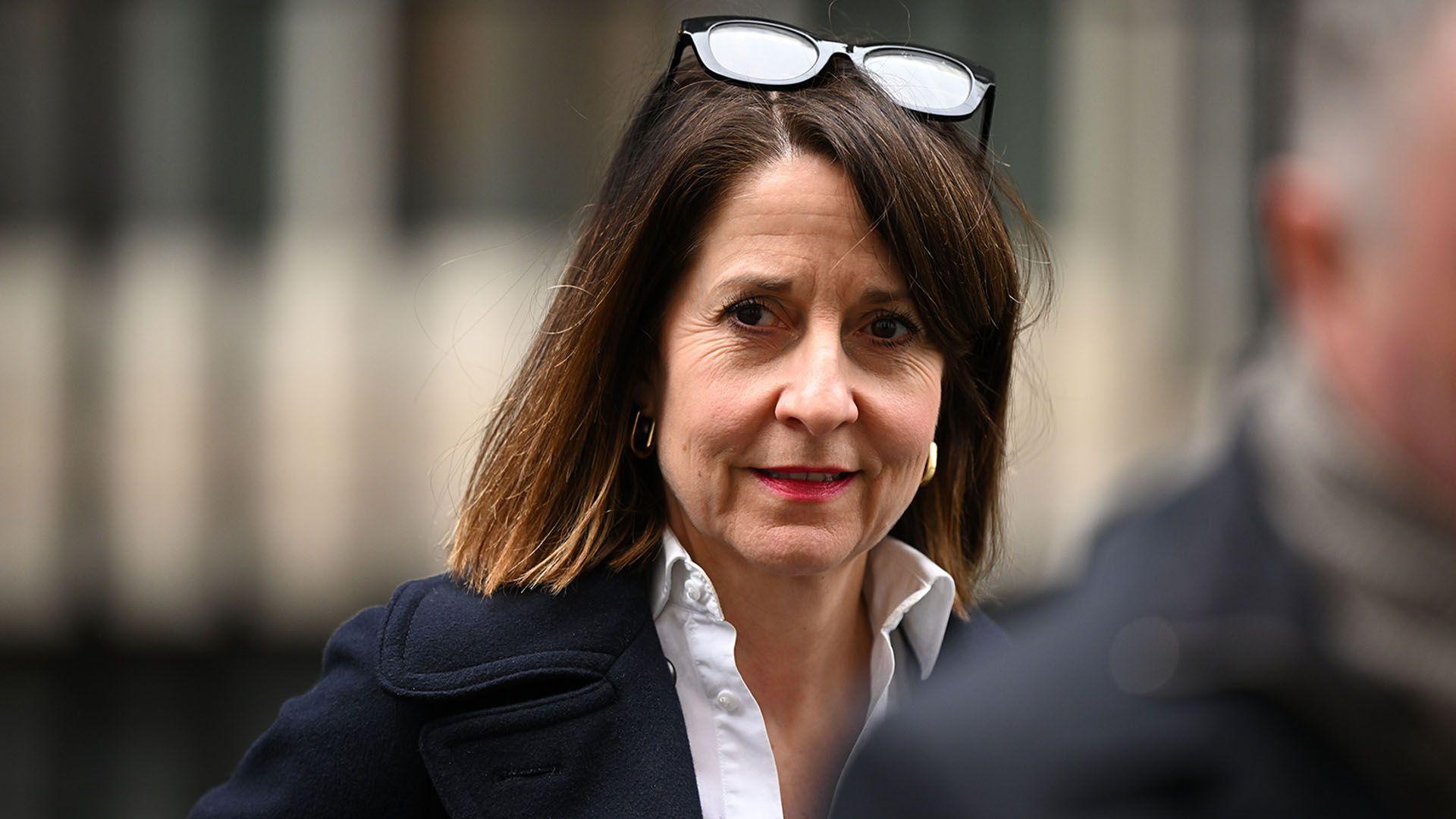 Head and shoulders shot of a woman wearing a white shirt and dark jacket. She has shoulder length brown hair and a pair of black glasses are resting on the top of her head.