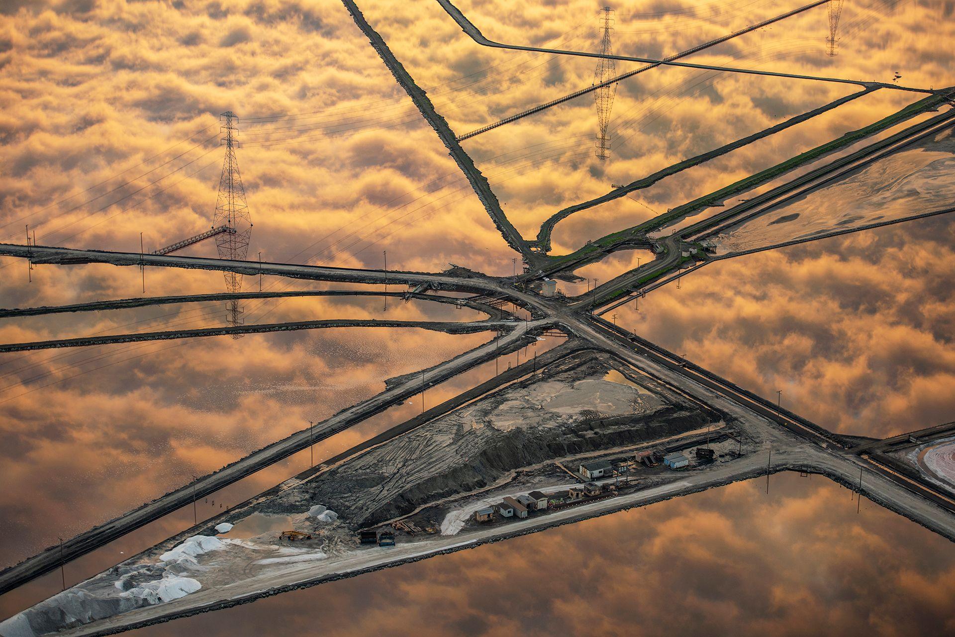 Golden-hued clouds are reflected perfectly in the grid-like pattern of salt ponds. The ponds stretch across the landscape, creating a patchwork of shimmering water and earth dividers.