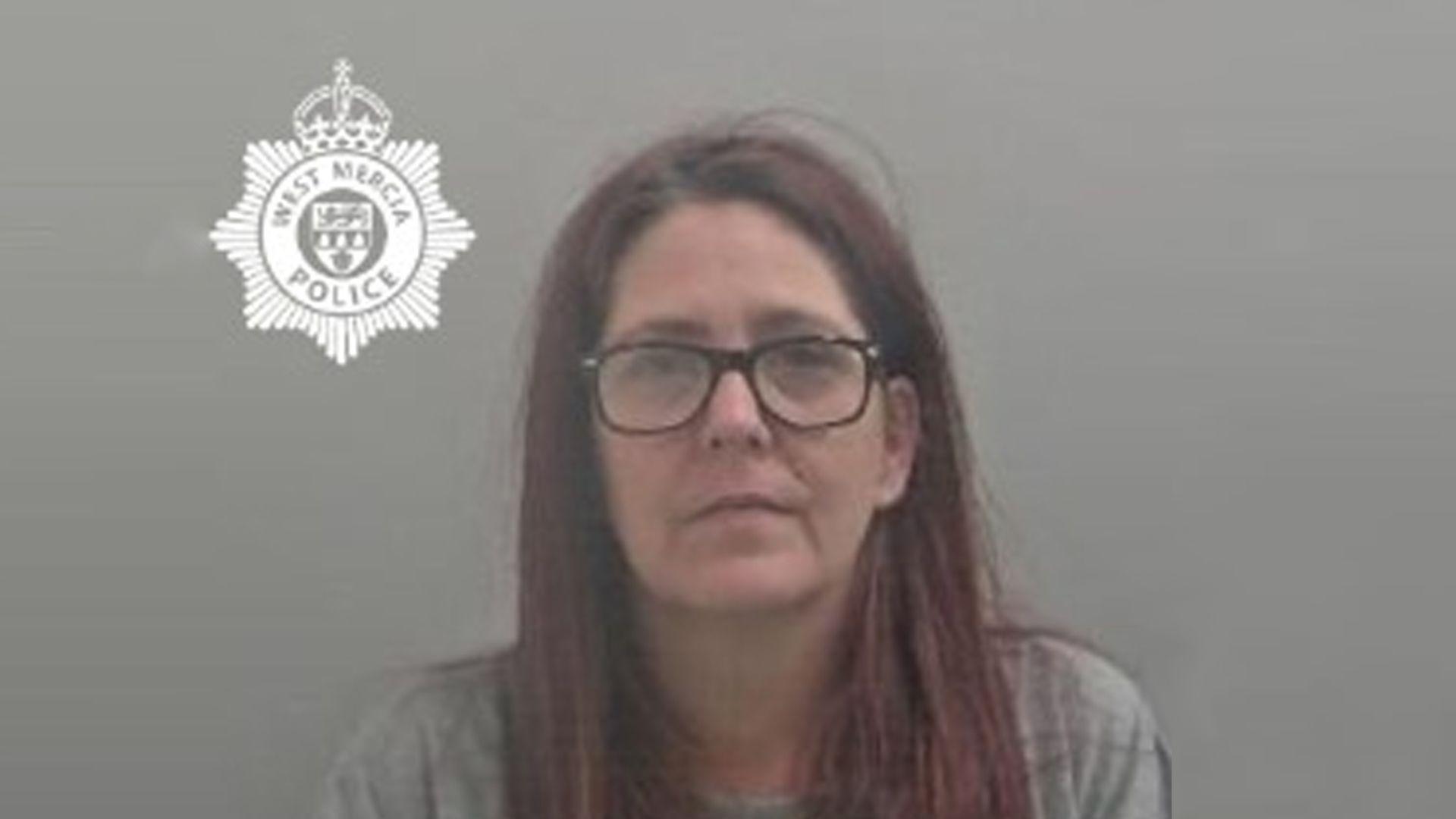 A woman's mug shot. She has purple-red hair and is wearing large circular glasses and a long sleeve grey jumper on.