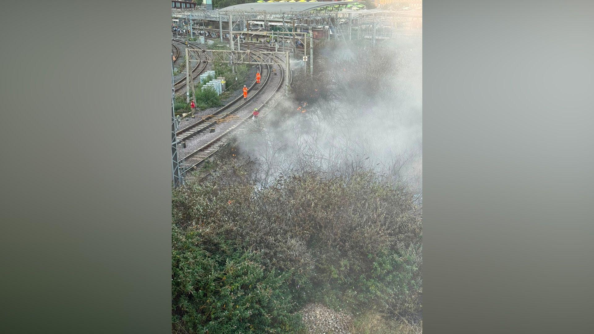Smoke pours from bushes close to a railway line in an urban area.