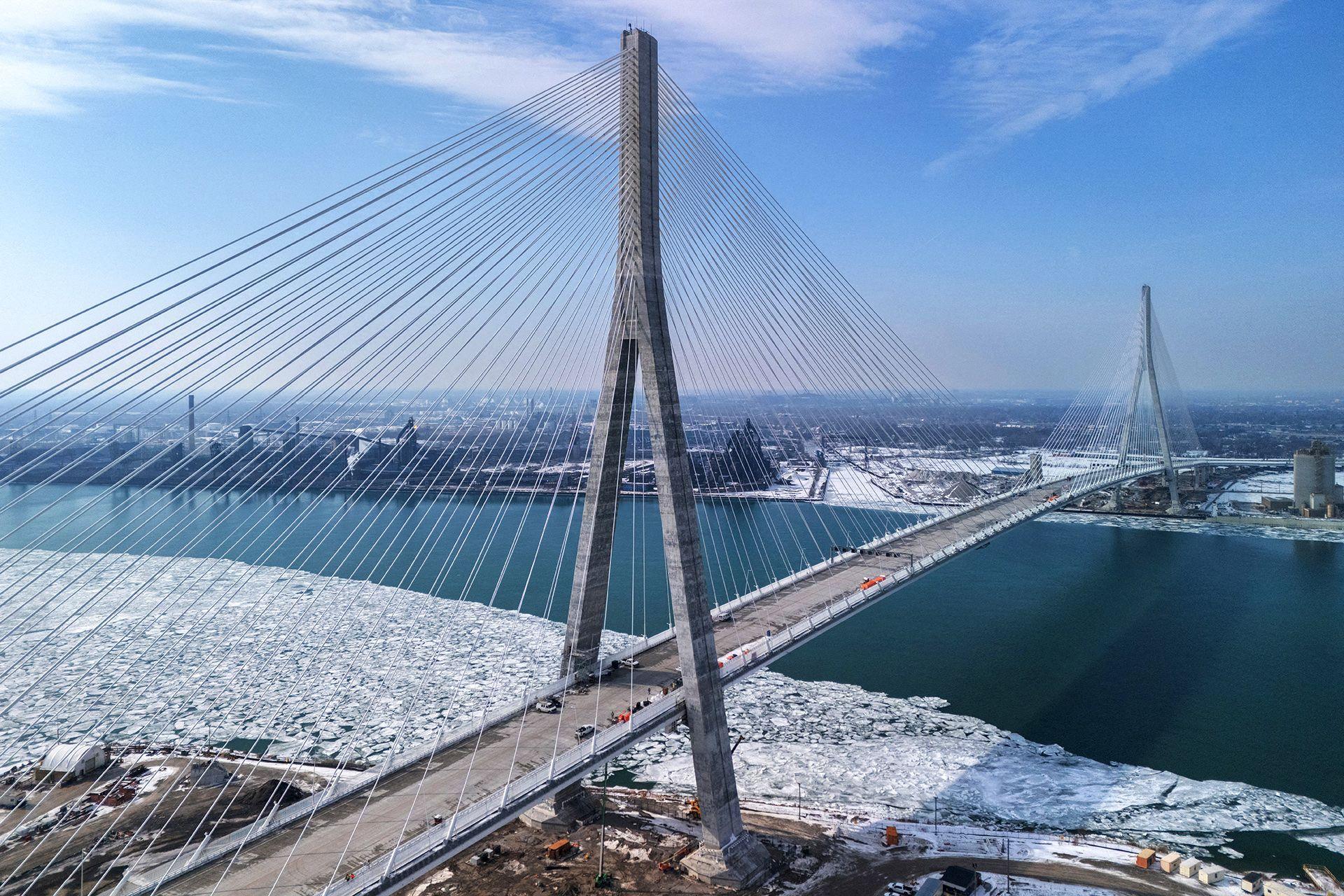The Gordie Howe International Bridge, a cable-stayed bridge featuring six traffic lanes, is seen in a drone image. The ground on either side is covered in snow, with unfrozen water flowing beneath. 