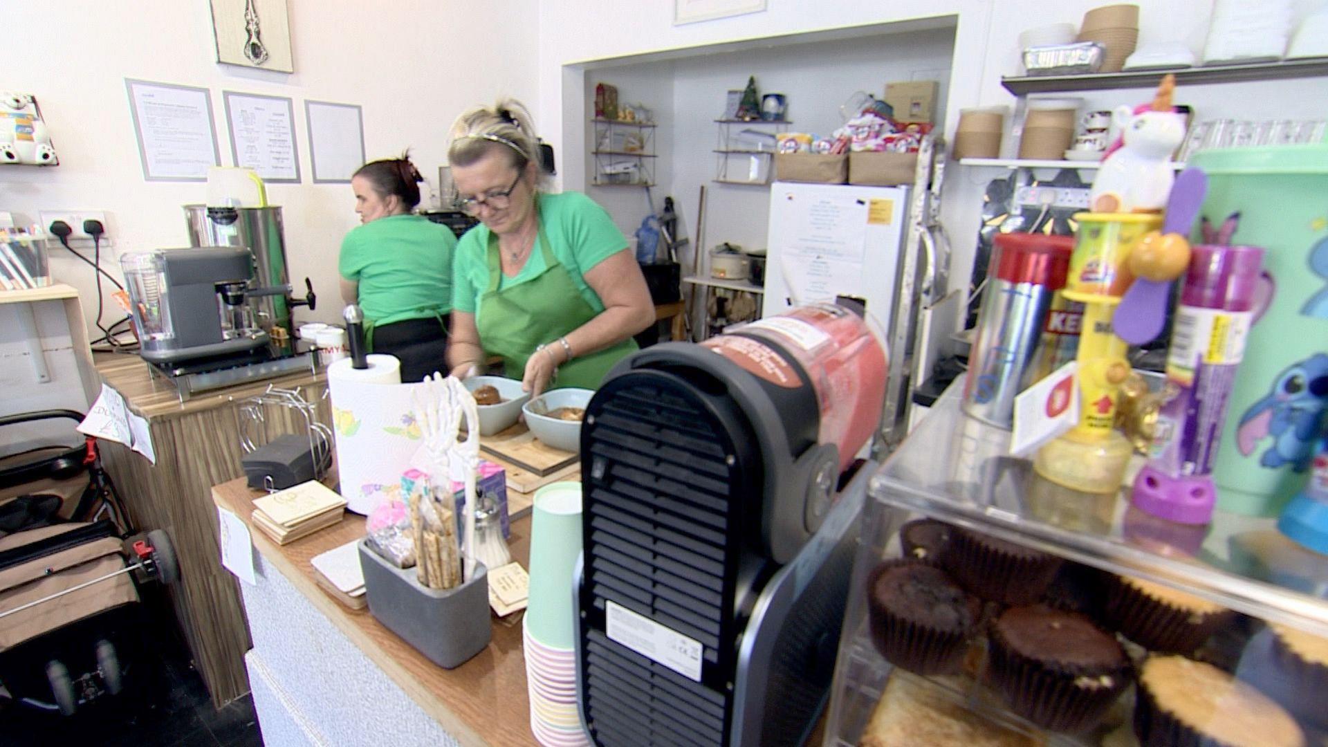 Inside a cafe with white walls and wooden worktops, Sam Elliott and Gemma Davis are wearing green t-shirts and aprons. Gemma Davis is standing by the sink with her back to the camera, and Sam Elliott is preparing two bowls of food. Surrounding them are various kitchen items including kitchen roll, paper cups, and coffee and slush machines.