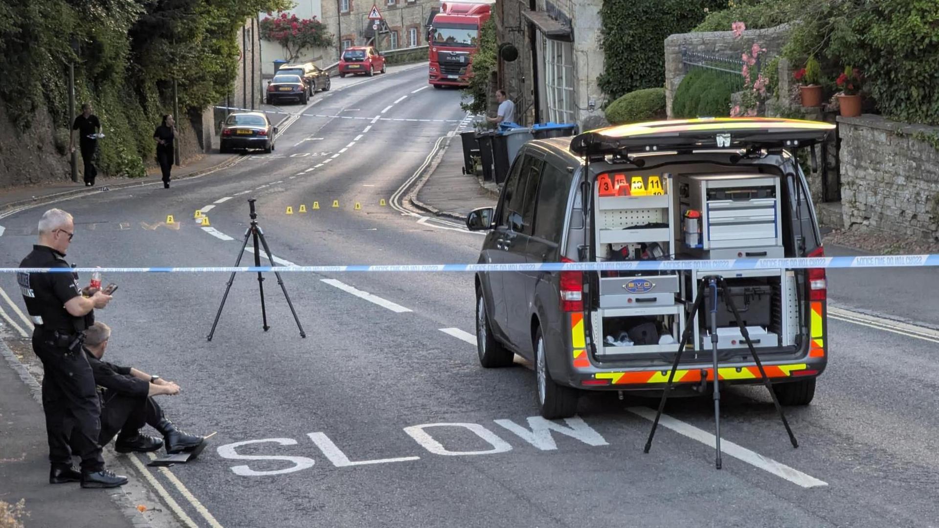 A police van on the main road in Woodstock. Blue tape surrounds the scene, with some numbered markers in the road. Police officers stand by.