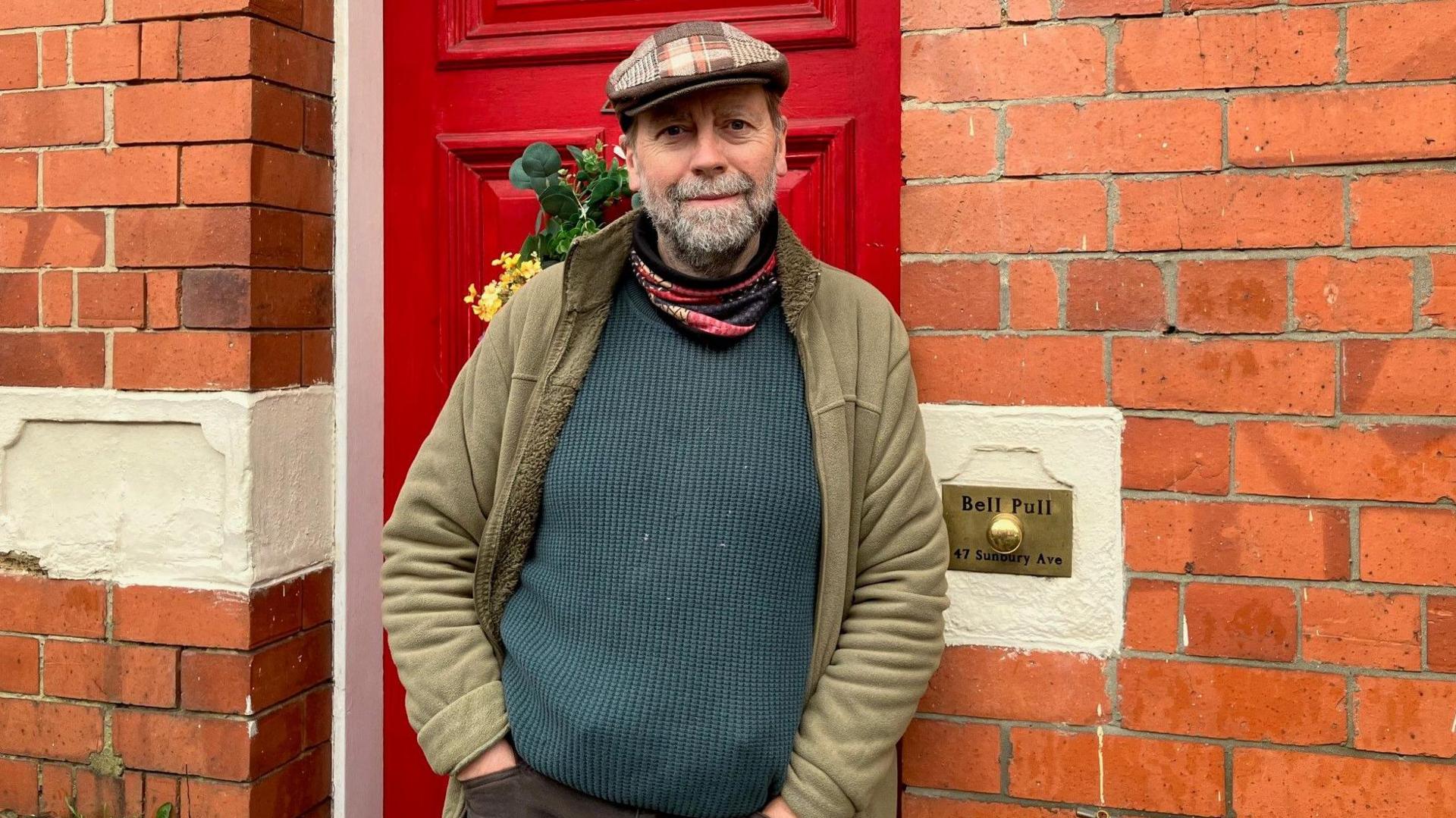 Christopher Wanche is standing on the doorstep of his redbrick house. Behind him is a red door with a wreath of flowers hanging off it and next to his elbow is an old-fashioned bell pull. He has a beard flecked with grey and is wearing a flat cap, a green coat and a dark green jumper.