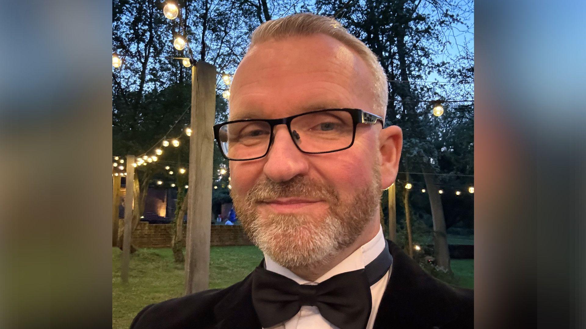 Mr Lait smiles at the camera while wearing a black suit with a bow tie and white shirt. He appears to be at a party or function. It is getting dark and there are strings of lights between poles behind him.