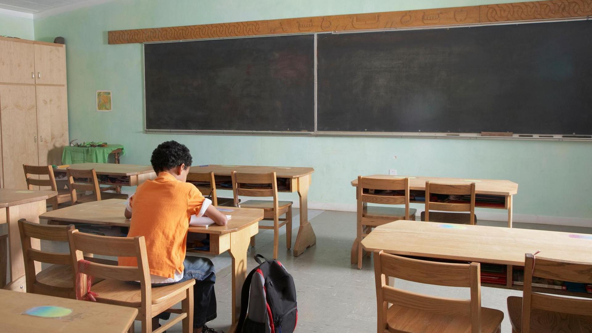 A child is sitting at a desk alone. There are five desks in the room. There is a blackboard on the front wall, the board is divided in two. The wall of the room is painted green. The child is wearing an orange top with jeans. Their backpack is black and is sitting beside them. There is also a large wooden cupboard in the background.
