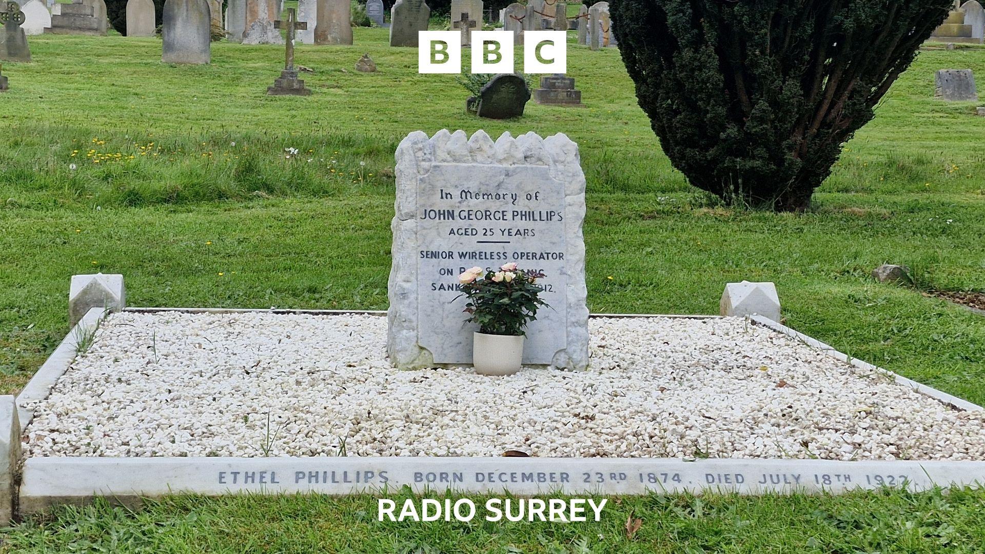 A small white gravestone sits on a square of white pebbles.  The gravestone reads 'In  memory of John George Phillips aged 25 years. Senior Wireless operator...' the rest of the wording is hidden behind a rose plant in a white pot.