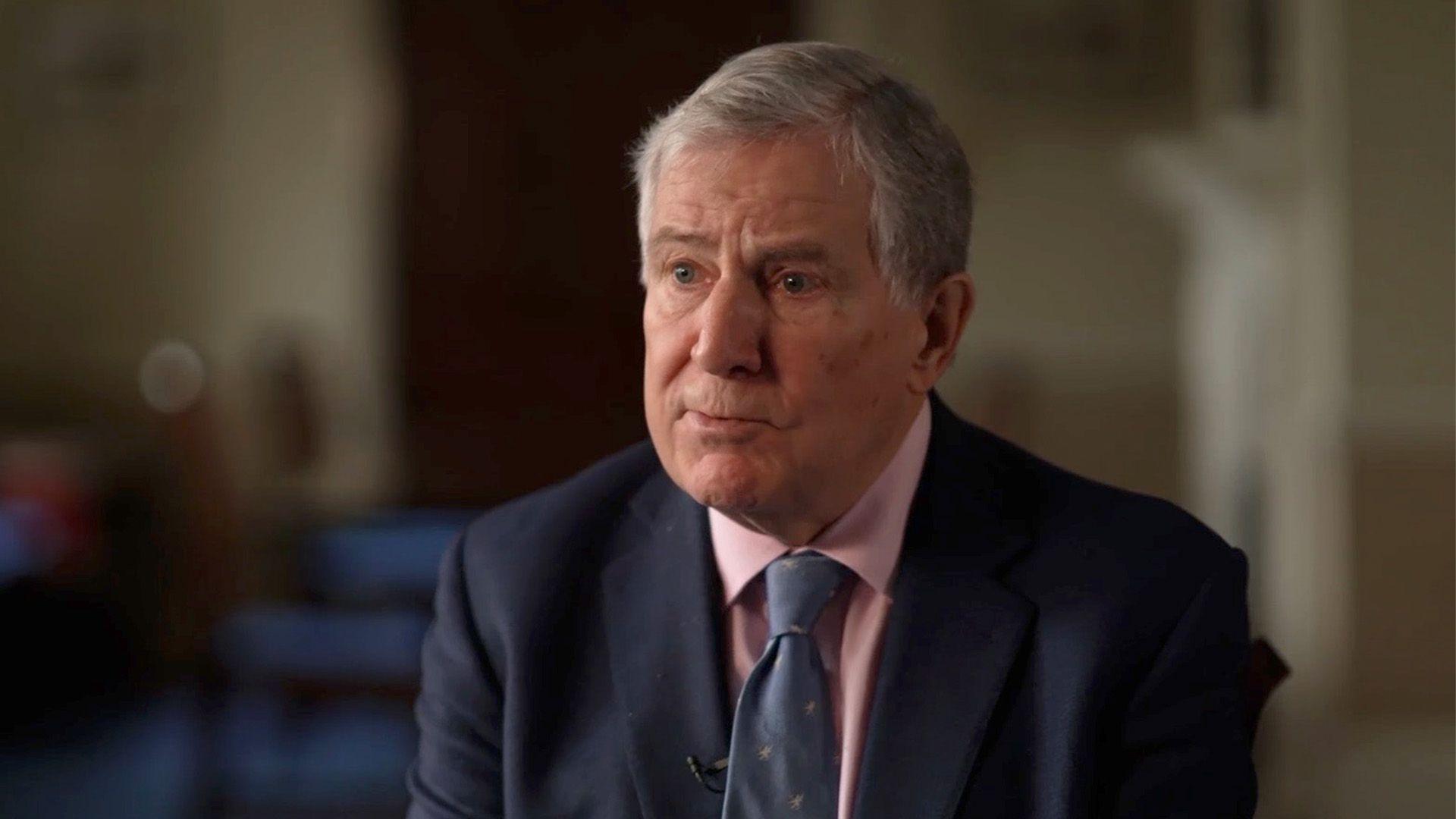 A head-and-shoulders portrait of Bruce Houlder, a white man with grey hair, wearing a navy suit, a pink shirt and a blue tie, looking to the side of the camera with the background out of focus