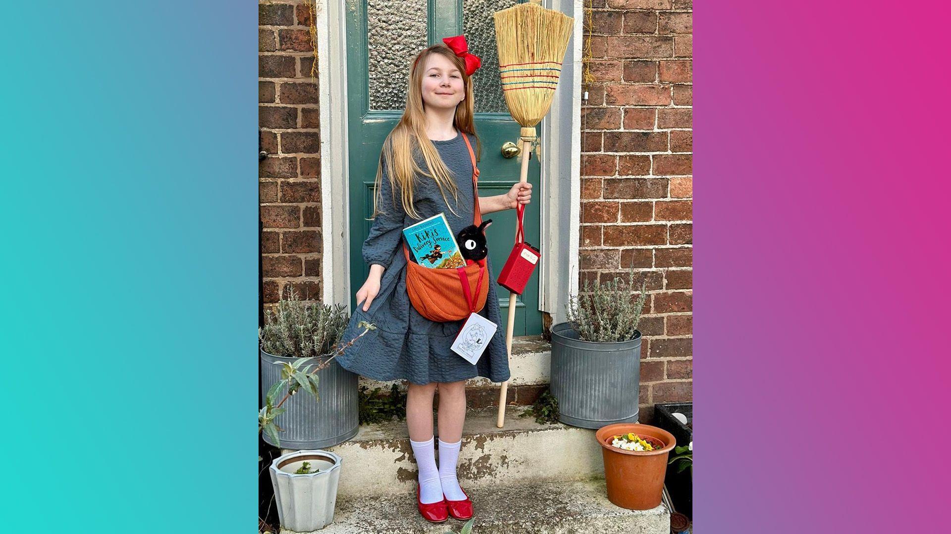 Scouty is standing in front of a front door. She is wearing a grey dress, white socks, red shoes and a red bow in her hair. She is holding a broomstick and in her orange bag, which goes across her body, there is a black cat plushie and the book. 