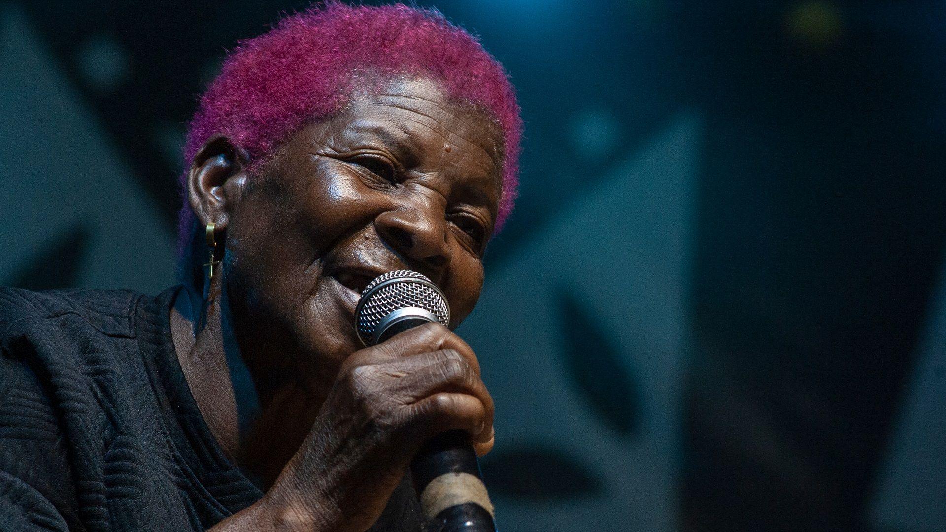 A woman in a black top with bright pink short hair sings into a microphone  during the Tumaini Festival at Dzaleka Refugee Camp in Dowa, Dowa, Malawi - Saturday 2 November 2024