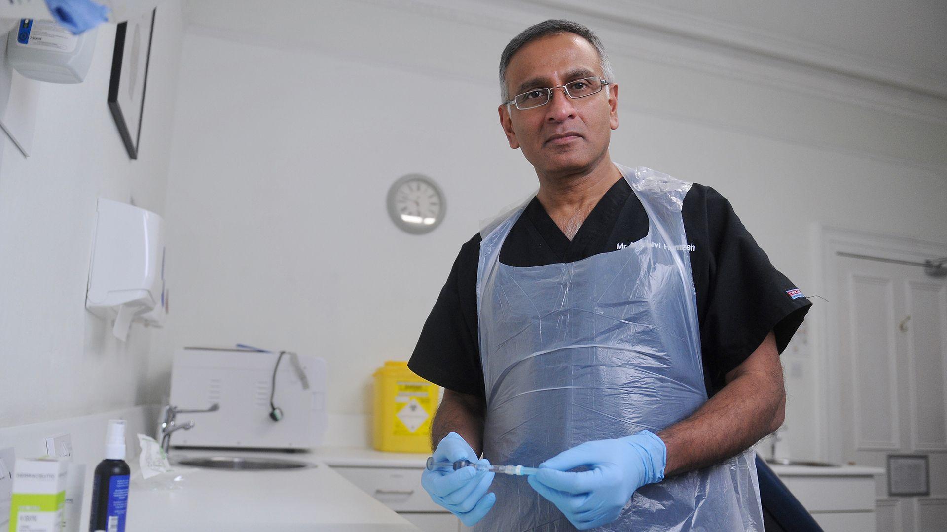 Dalvi Humzah in his white clinical treatment room, holding a syringe and wearing a plastic apron and rubber gloves. In the background a sink, sharps bin and disinfectant dispensers - the kind of hygienic resources that are standard in a clinical setting.  