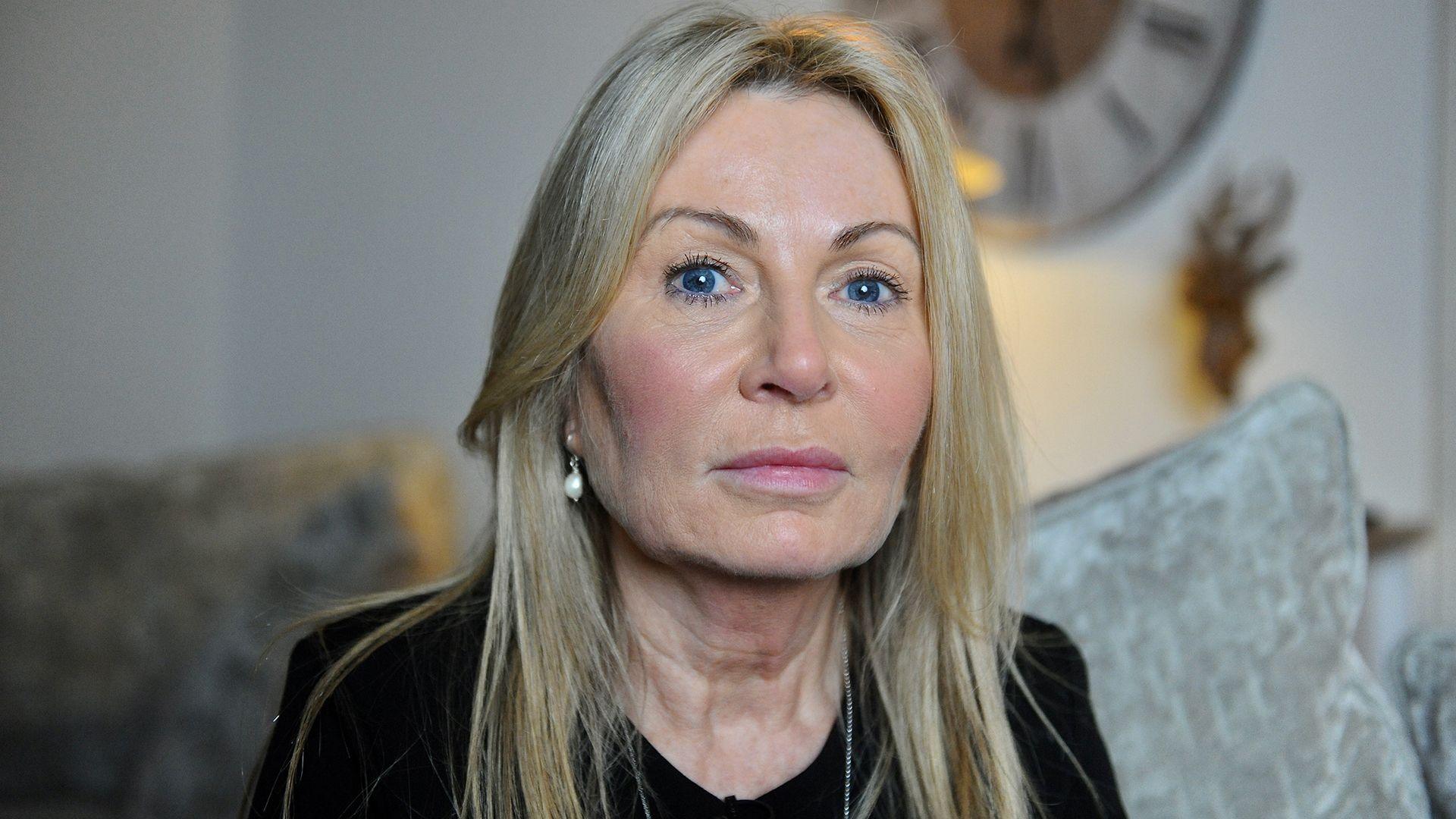 Janet Taylor, with long blonde hair and blue eyes, wearing pearl earrings, sitting on the grey couch in her living room, looking straight at the camera 