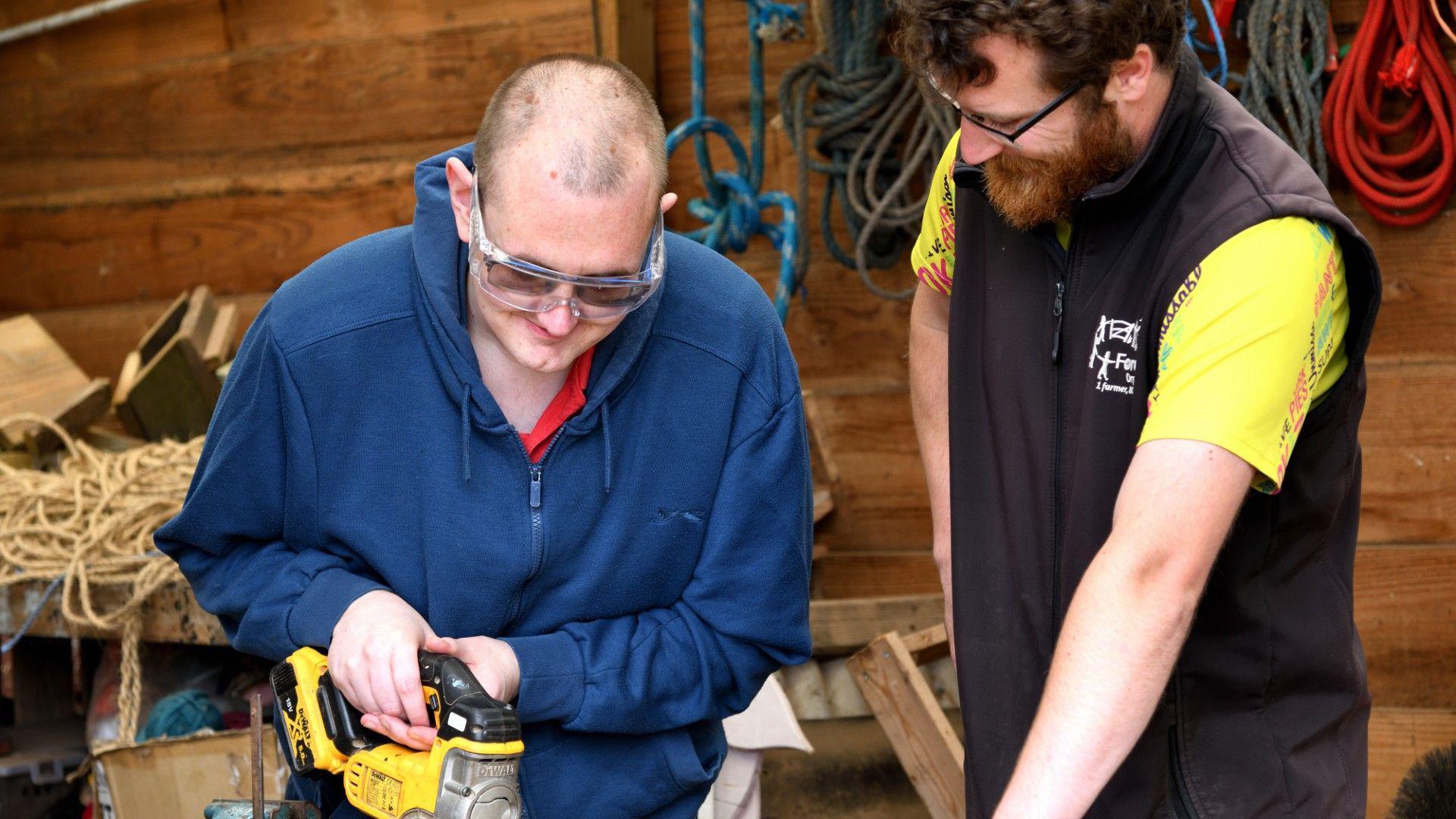 On the left, there is a man with a shaved head. He is wearing see-through safety goggles and dark glasses. He has a blue Slazenger cardigan with a zipper with a red polo shirt underneath. He is holding a yellow power tool. On the right, there is a man with brown curly hair, a ginger-brown beard and blue glasses. He is wearing a yellow tshirt with a black sleeveless vest.