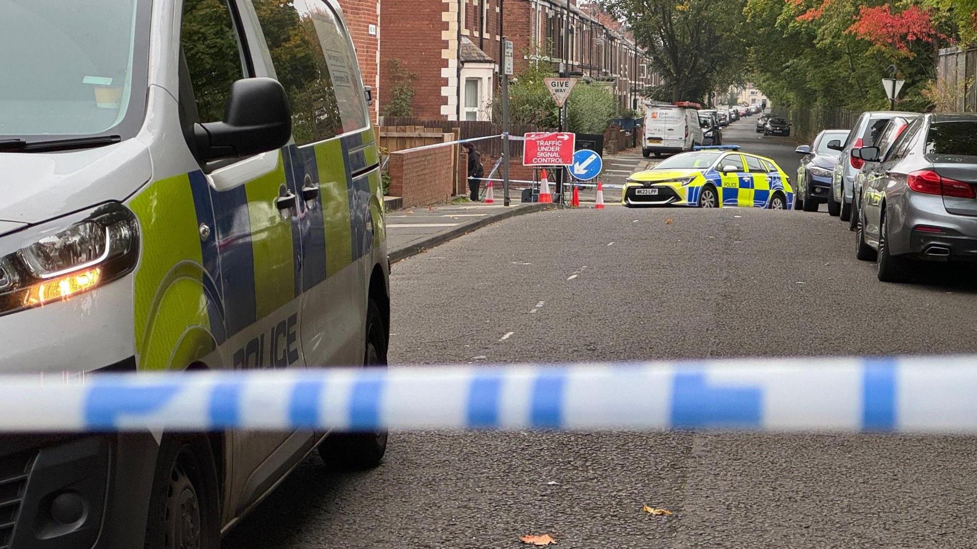 A police car next to a cordoned off street. Further down the road is another police vehicle. 