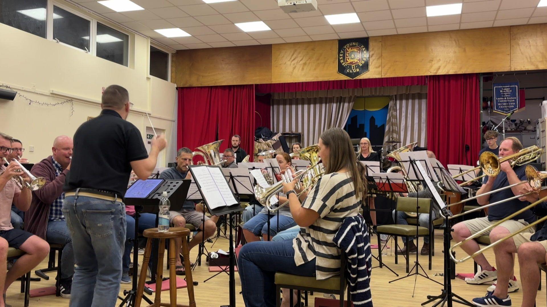 A man in a black polo shirt and jeans conducts a brass band, made up of male and female musicians. There is a trumpet and some tubas in the foreground, and a French horn further back.