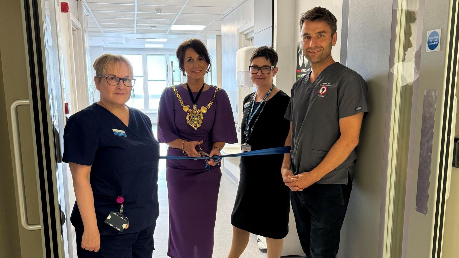 Ms Dunn wears a purple dress as she holds scissors to cut the blue ribbon stretched between two doors. Three trust staff stand next to her, including Dr Ball on the very right. 