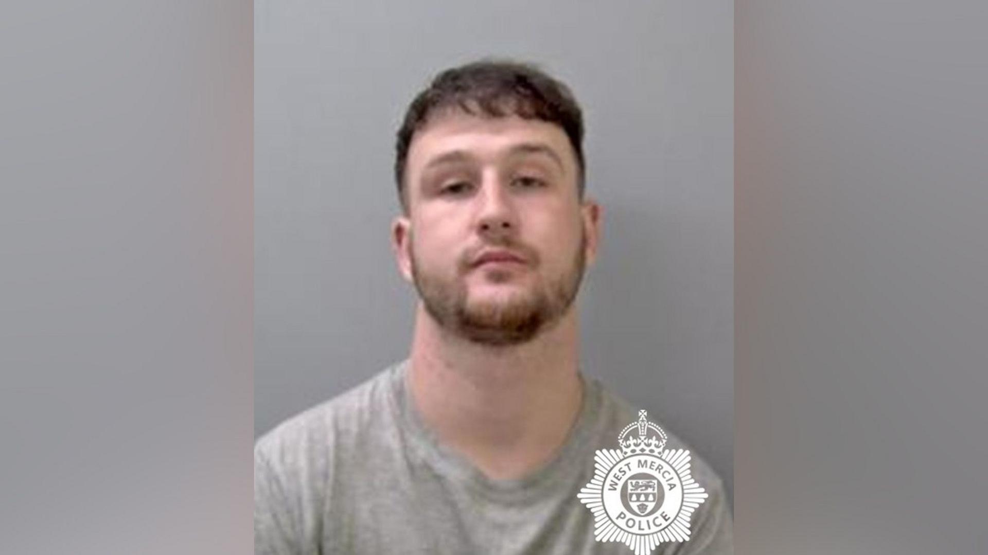 A mugshot of a man with brown beard and fringe. The picture is against a grey background and the man has a serious expression.