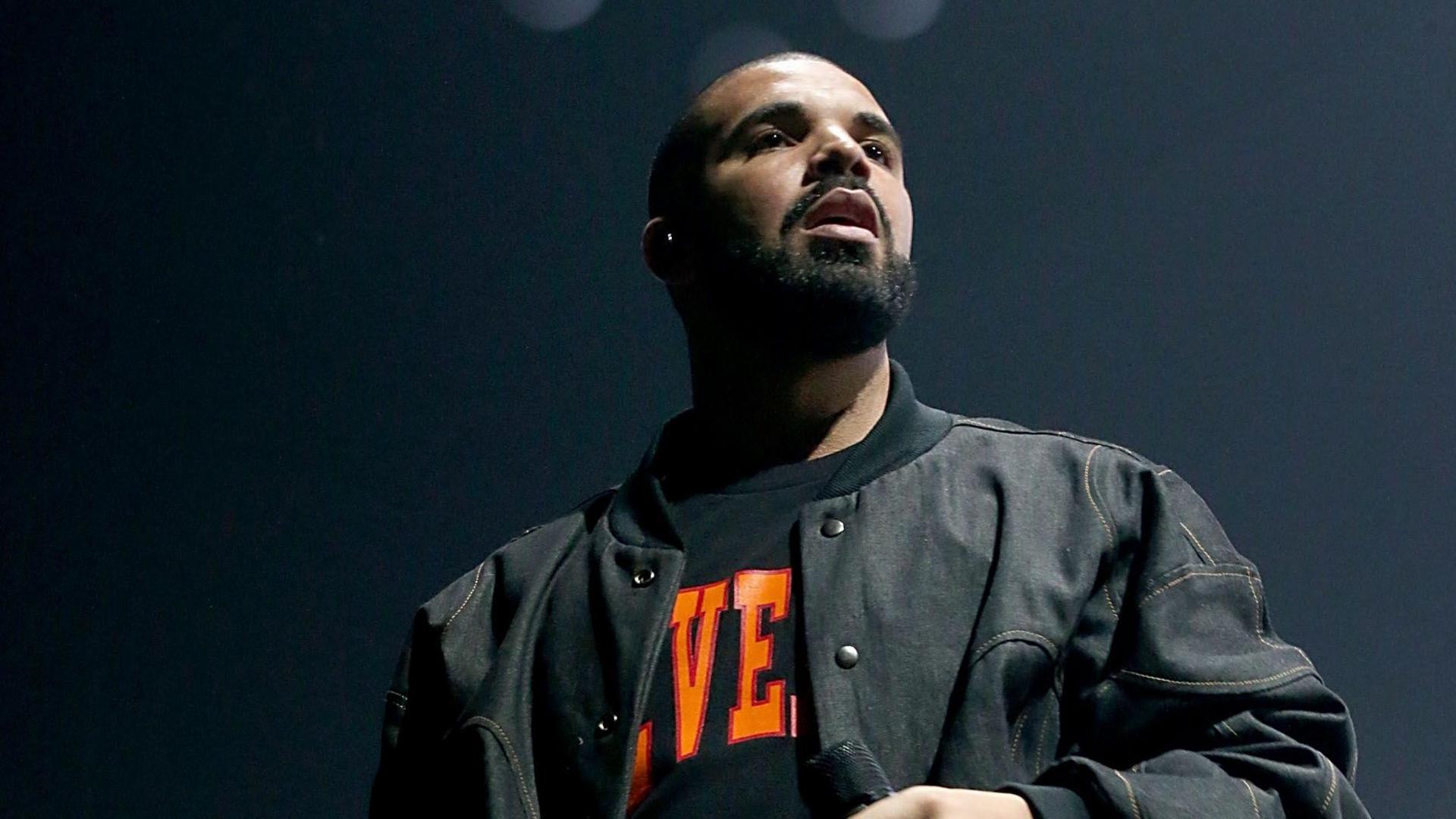 Drake performs on stage in a black jacket with a branded t-shirt partially on show