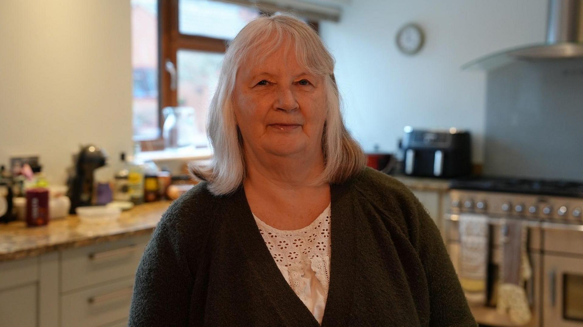 Ruth Matthews standing in her kitchen. She has grey shoulder length hair and a fringe, and wears a white lacy blouse with a green cardigan over it. In the  background is a Shaker style kitchen with a large range cooker, and a kitchen window.