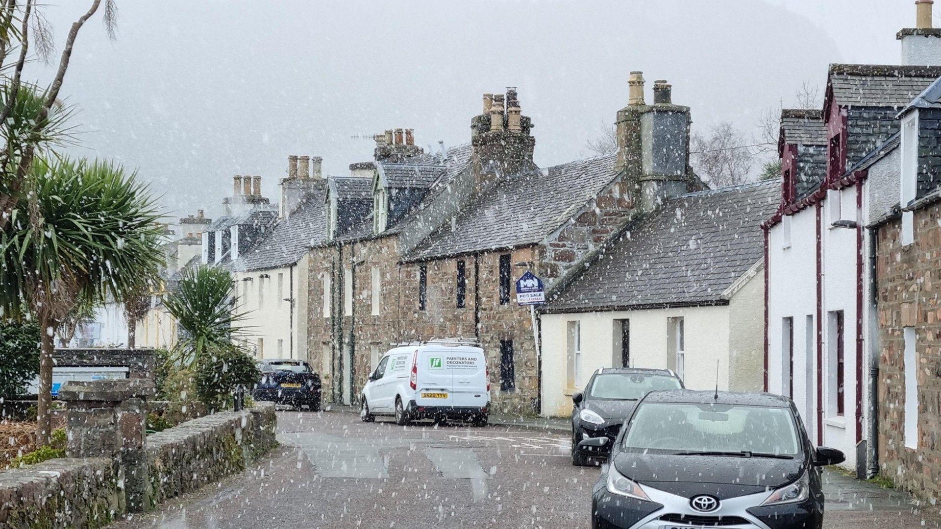 Village scene showing road and parked cars with snow falling