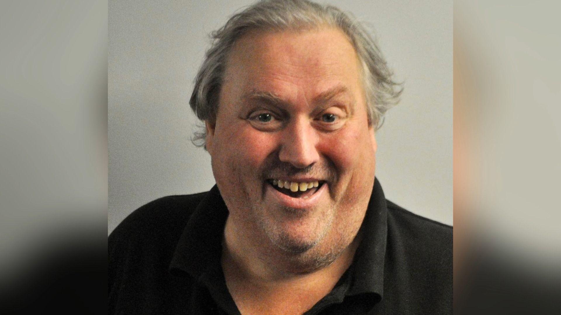Paul Brammer, a man with grey hair is smiling. He is wearing a black polo shirt, and is stood against a pale coloured backdrop.
