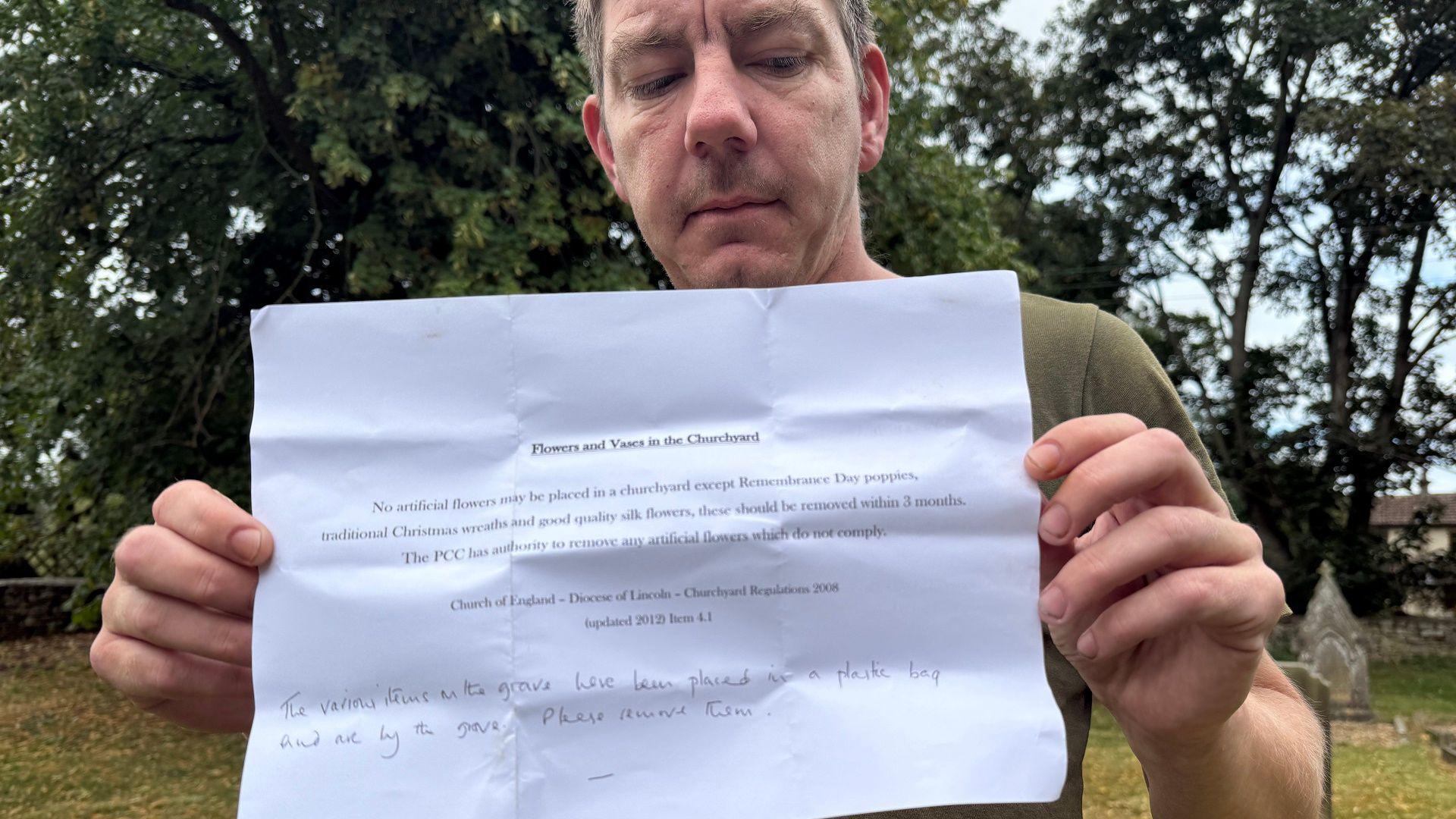 A man with grey hair and wearing a green t-shirt holds up a letter. He is standing in a grass covered graveyard surrounded by trees.