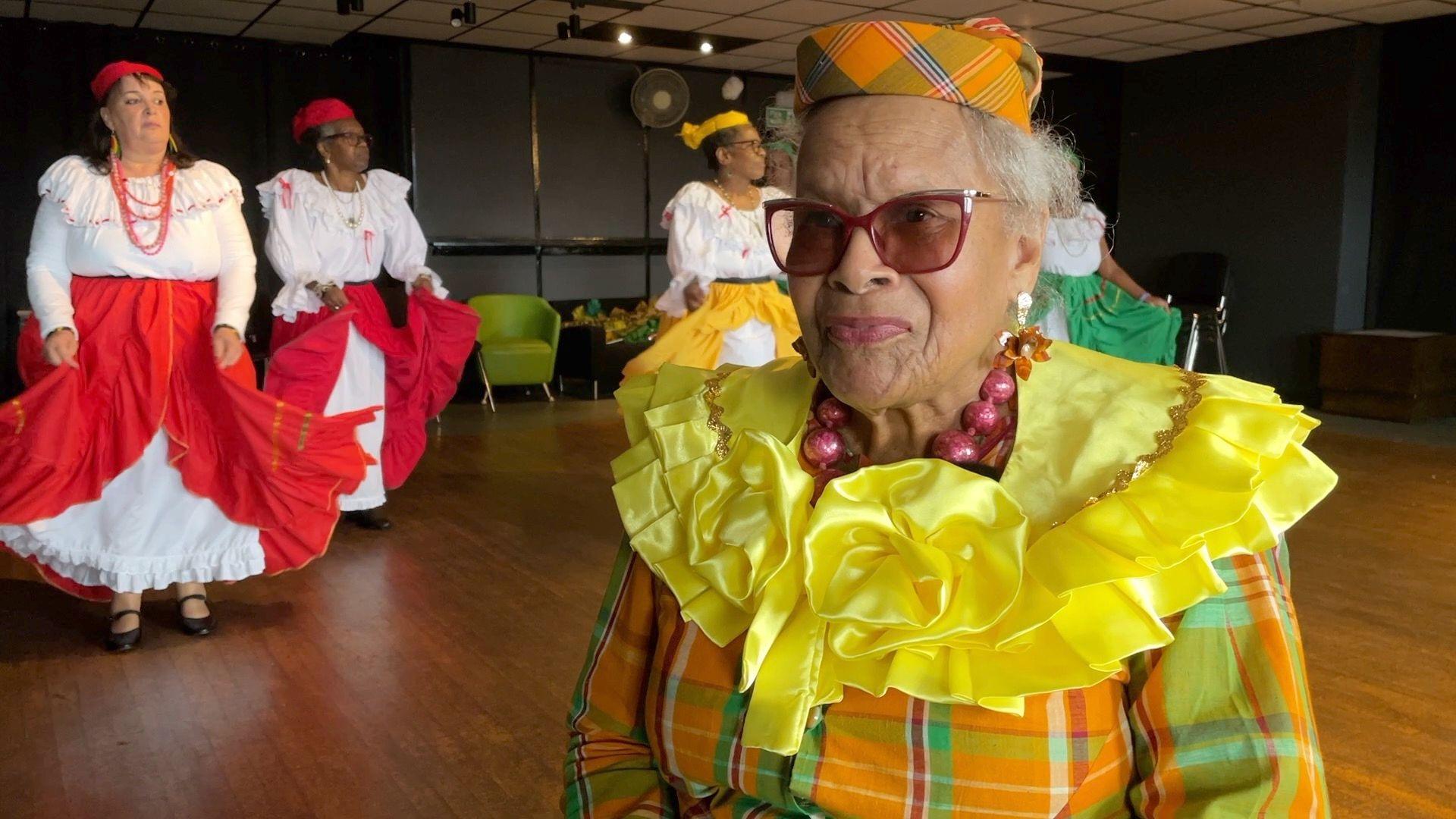 Patty Scott wears a traditional Quadrille dance costume of vibrant yellow, orange and green hues. Performers are dancing in the background.