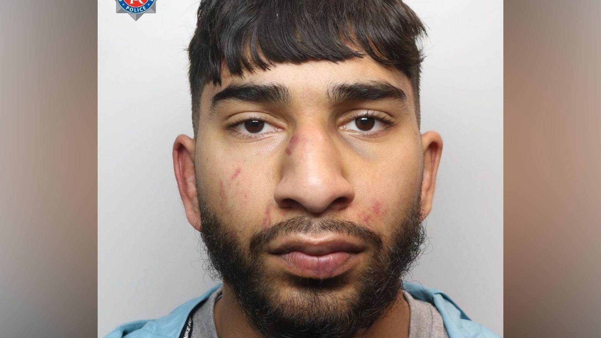 A mugshot of an Asian man in his early 20s, who has black hair cut into a fringe and black facial hair. He has red marks to his face  and is looking into the camera with a blank expression. 