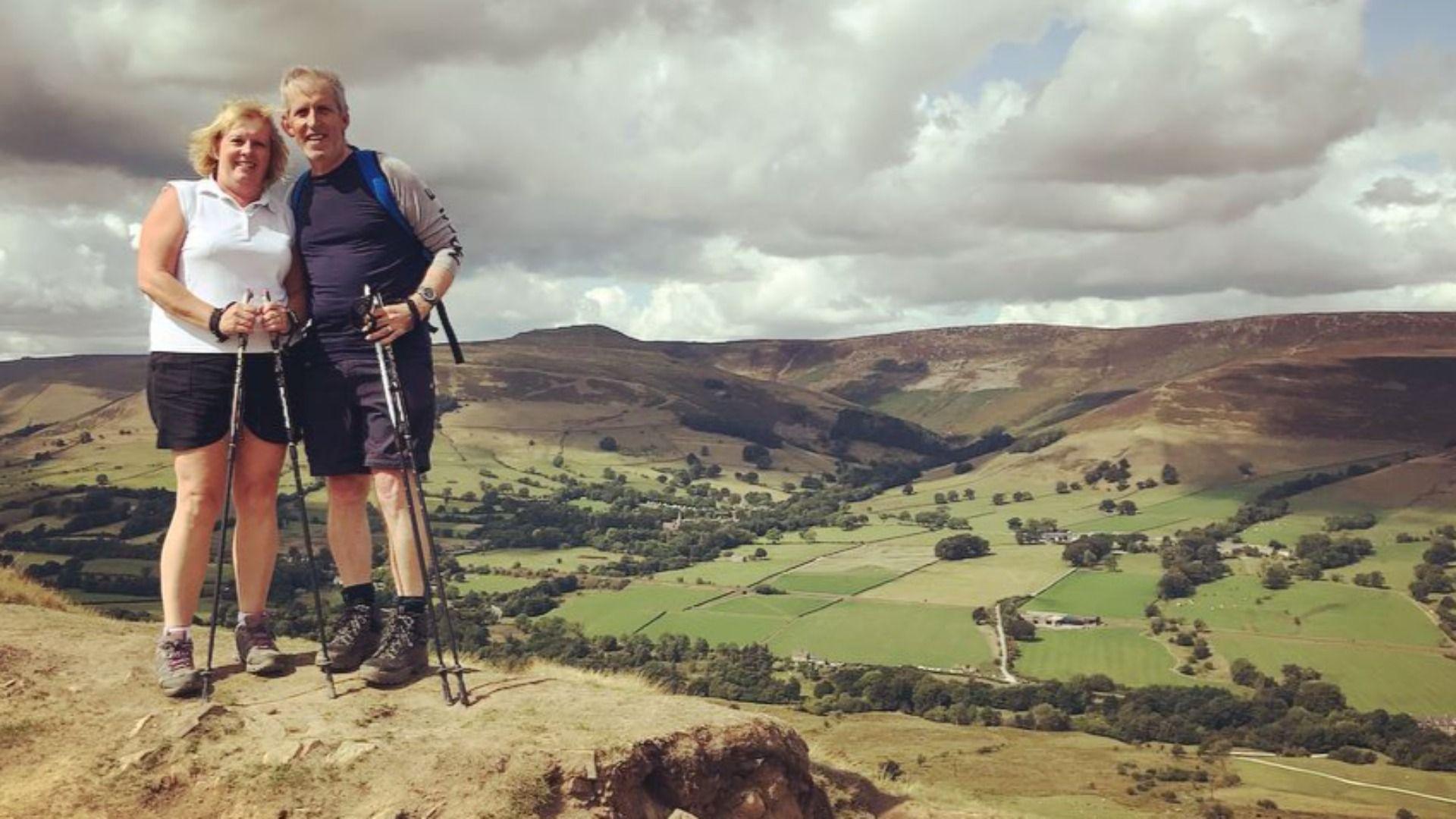 Pete and Pauline Turfrey standing together on a high grassy verge overlooking a valley in the landscape with various fields, trees and mountains. They are both wearing hiking boots, black shorts, and holding thin Nordic walking poles, smiling at the camera. The sky is cloudy with spots of blue peeking through.