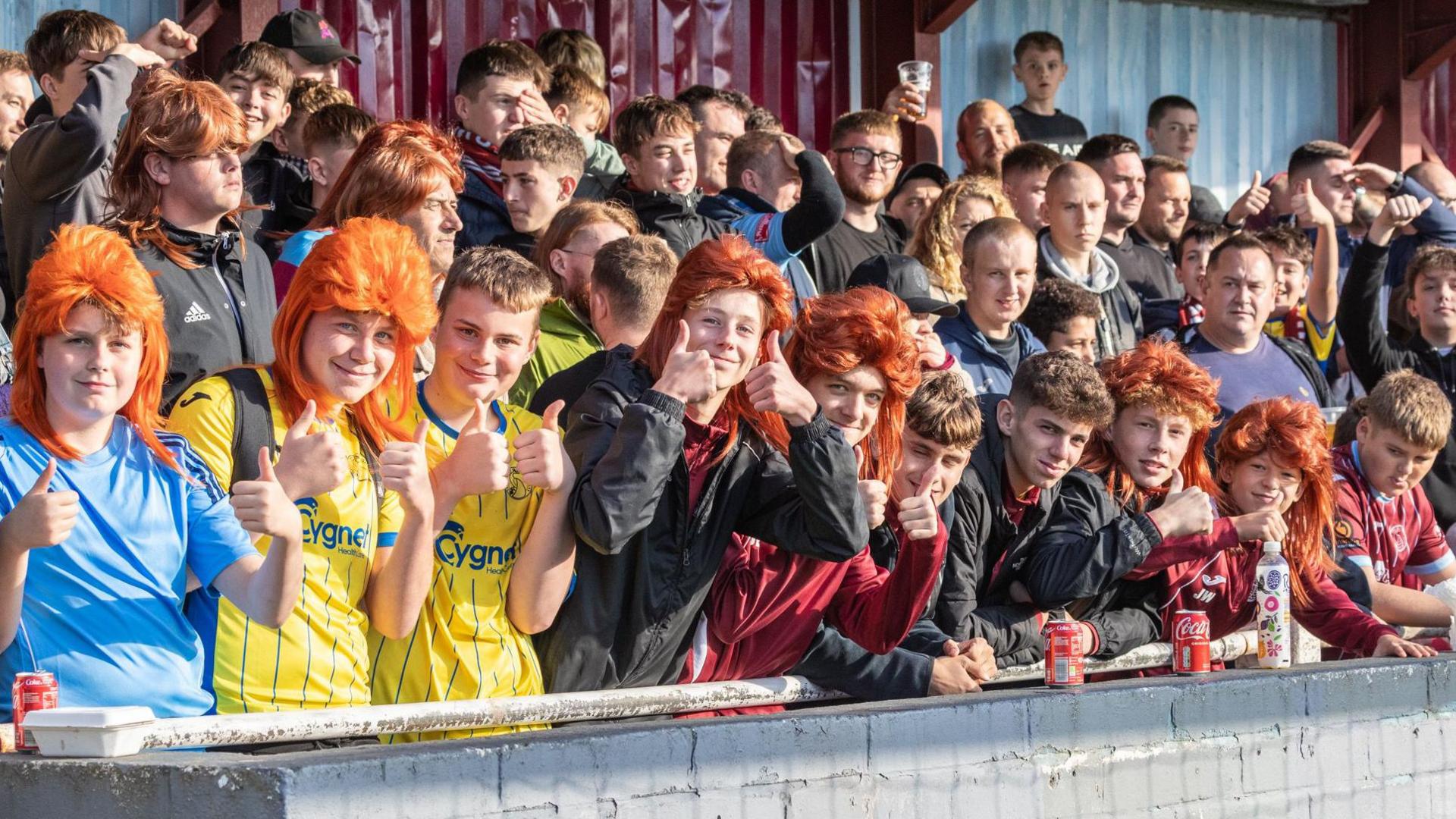 Football fans wearing orange mullet wigs. They are in the stands and wearing football merch.