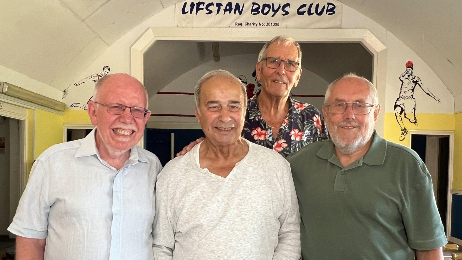 Four former members of the Lifstan Boys Club stand in front of a stage which bears the club's name. There is a domed white painted roof curving over the stage. The men are in their late 70s and early 80s. One on the left wears a light blue shirt and has a white goatee beard. Second from left is a man wearing a white open necked shirt. At the back is a taller man wearing a dark floral pattern shirt and glasses, and on the right end is a man wearing a dark green polo shirt, glasses and has a grey beard. All four men are smiling at the camera