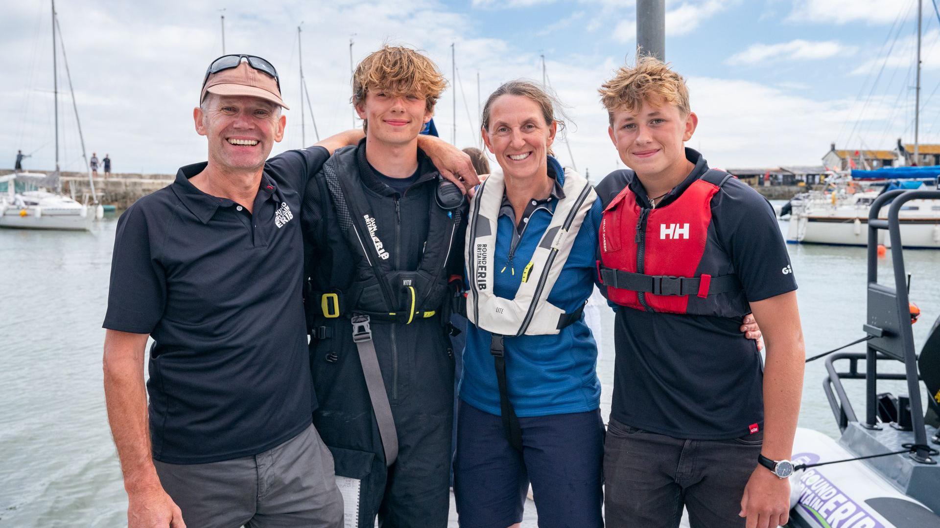 Harry Besley standing with Jacqui Besley, Henry Cietak, and another crew member, they all have their arms around each other and are standing in Lyme Regis harbour, with sailboats visible in the background. 