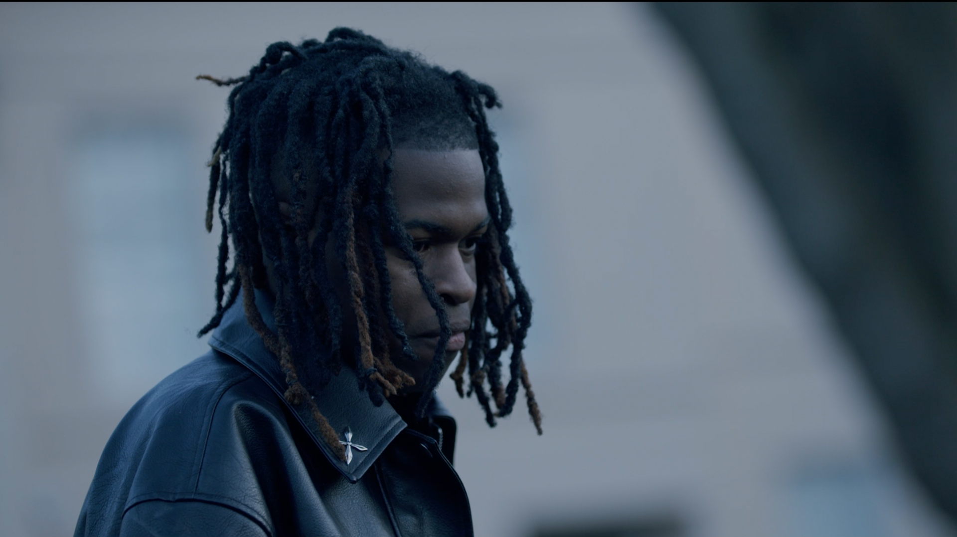 Jordan Adetunji. He has shoulder-length dark hair, wearing a black leather jacket. He is looking to his side.