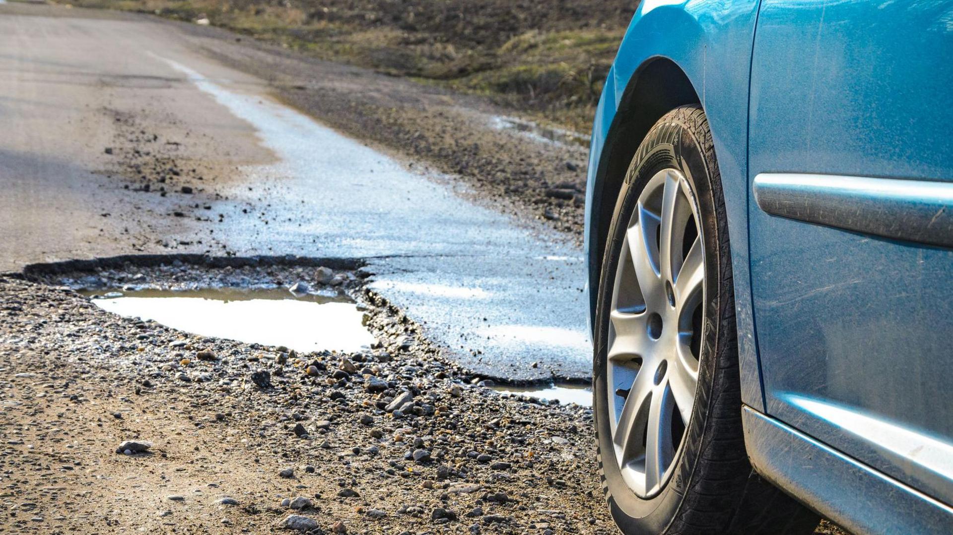 A blue car approaches a large pothole in a road