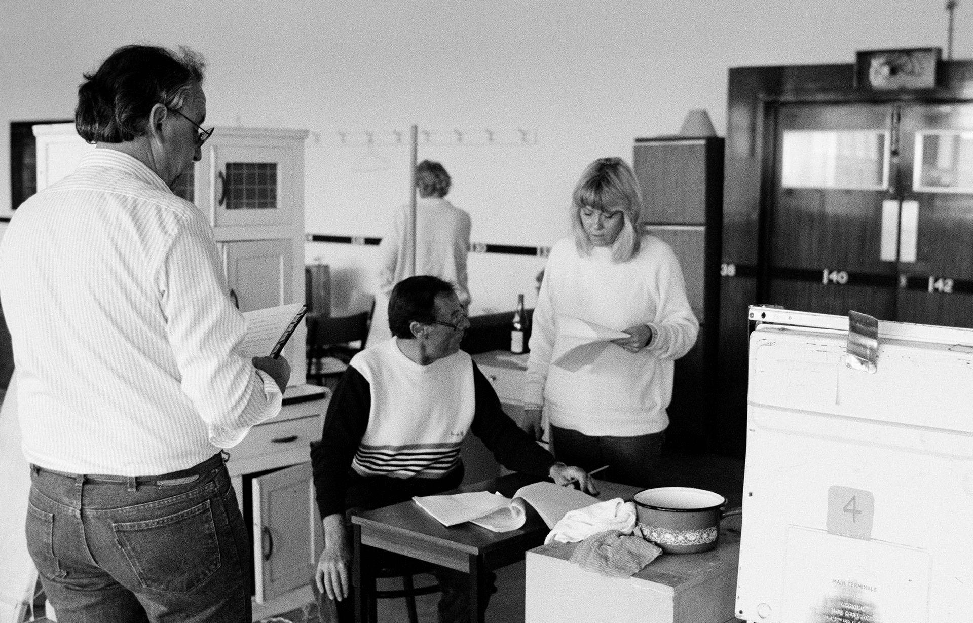 a black and white photo of wendy richard and bill treacher reading from their scripts amid various kitchen furniture props