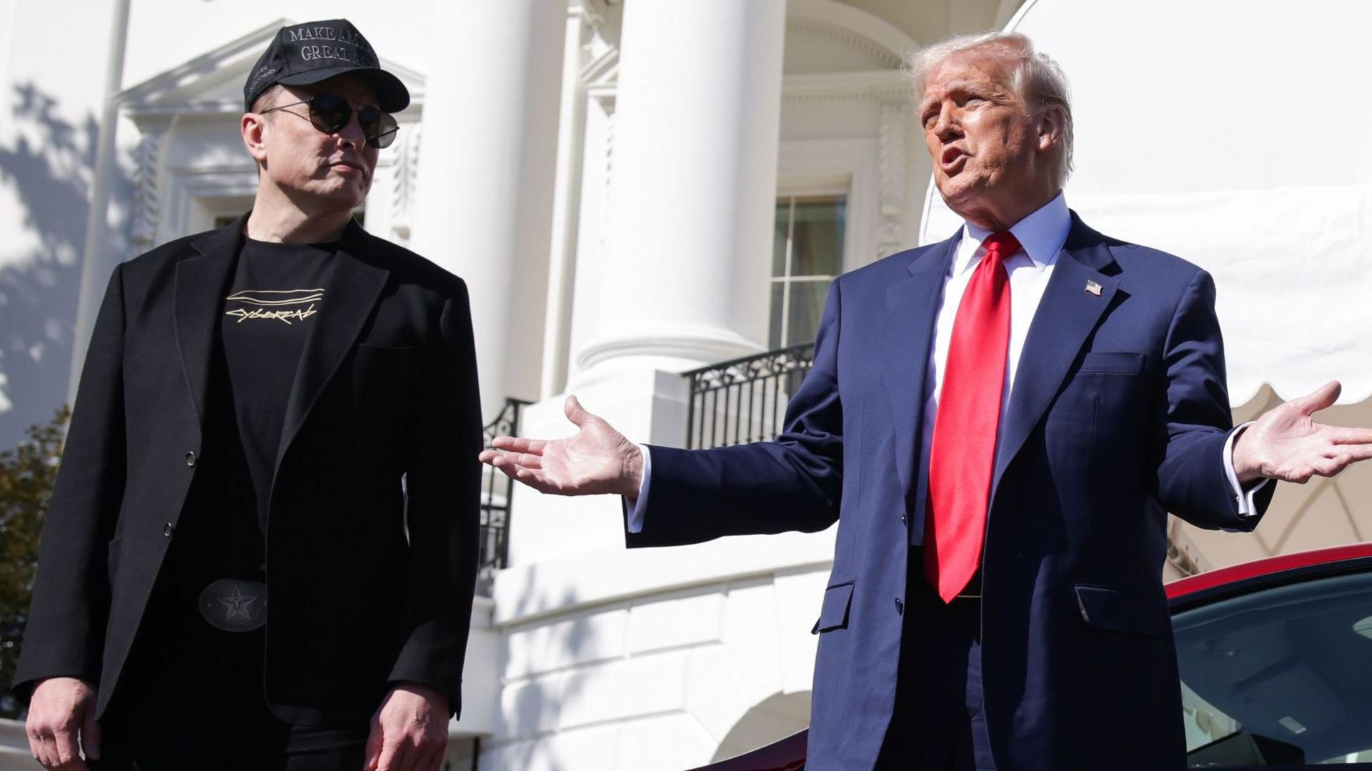 Elon Musk stands next to Donald Trump outside the White House as Trump gestures while speaking to reporters next to a red Tesla on 11 March