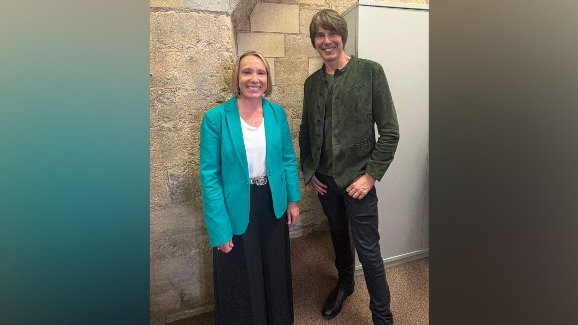 On the left, there is a woman in a turquoise-teal blazer, white t-shirt and black trousers. She has blonde ear-length hair and is smiling. On her right, there is a man with black jeans, black trousers and a dark grey jacket. He also has brown hair brushed to his left.