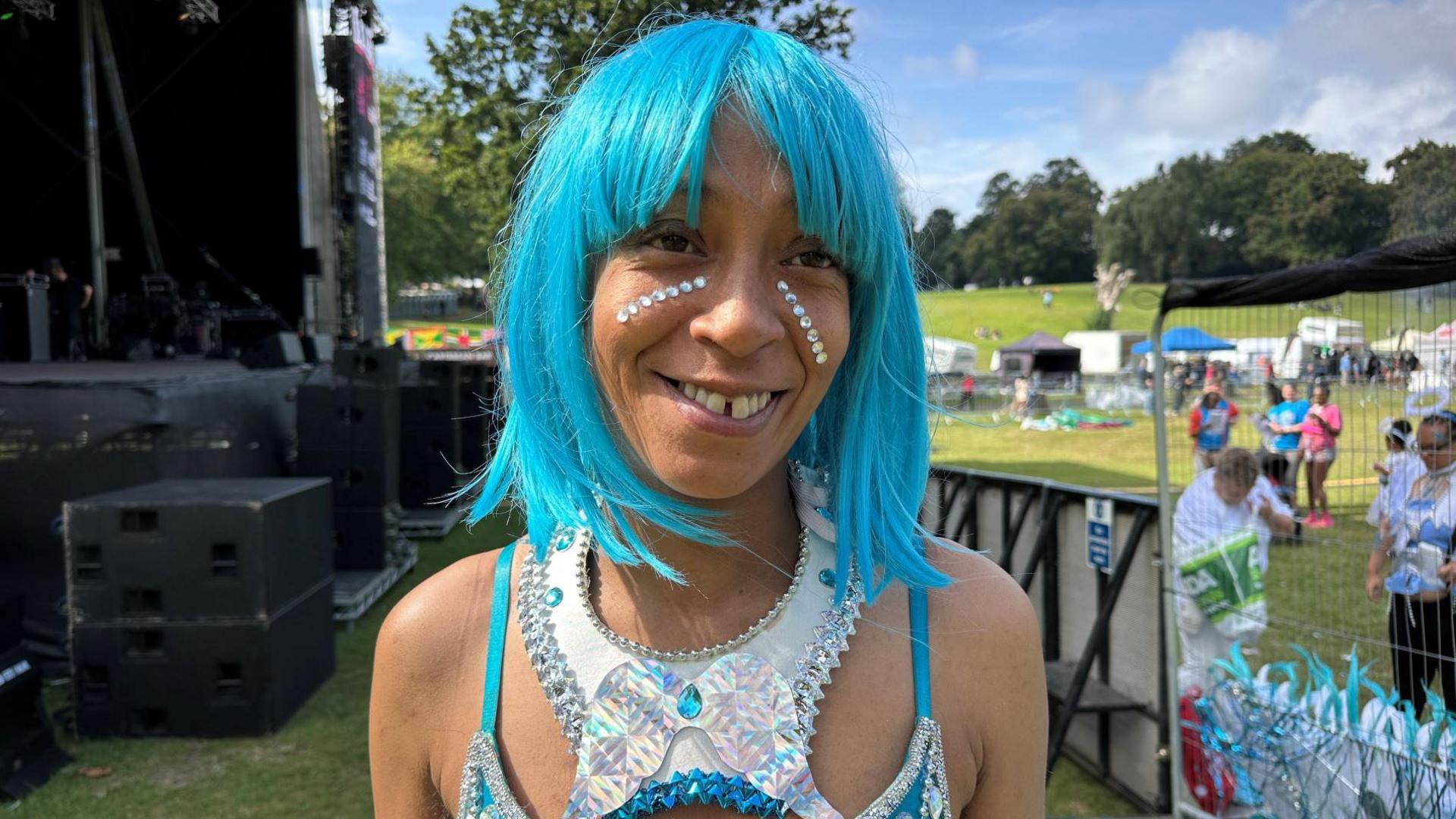 A smiling woman wears a turquoise wig and stick-on gems along her cheek which match her sparkling outfit.