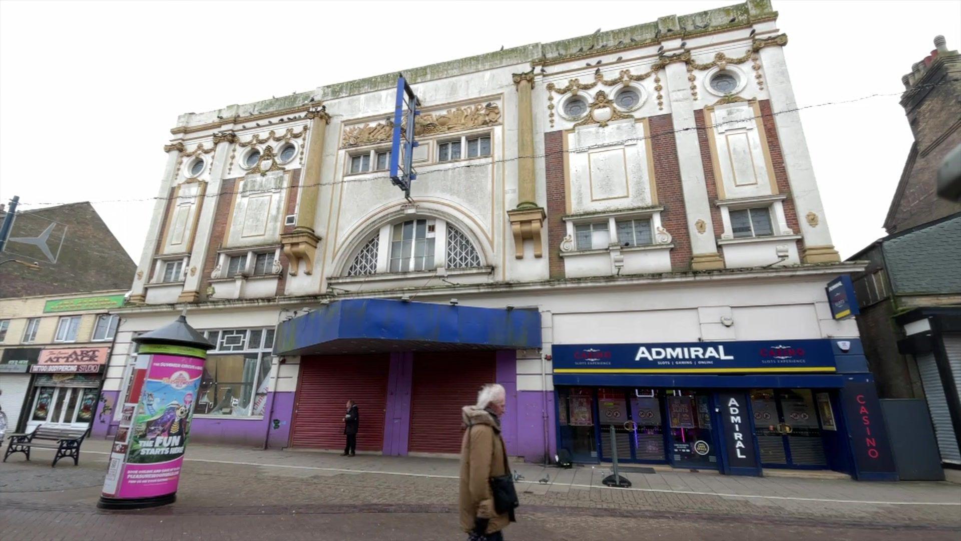 The Victorian looking theatre building can be seen on the high street in Great Yarmouth. The red shutters are down. It looks run down. There is a casino shop front on the ground floor. A person with white hair and a coat is walking in the foreground