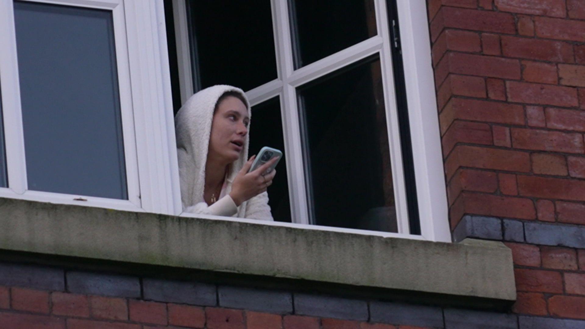 A woman with a furry hooded pyjama top leans out of a window holding her mobile phone in front of her face