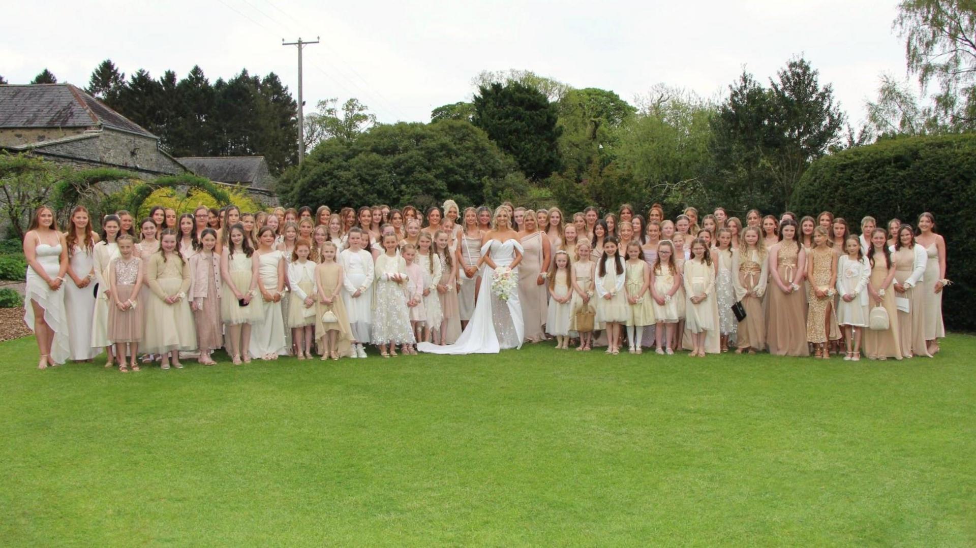 A wide shot photo of a bride, with blonde hair, a white dress with an off the shoulder detail carrying a bouquet, stands in the middle of a large group of women and young girls in the front row, wearing a mixture of champagne, cream, white and tan dresses. The women are standing in the middle of a large garden, with tall trees and a leafy tunnel to the left and are looking at the camera.