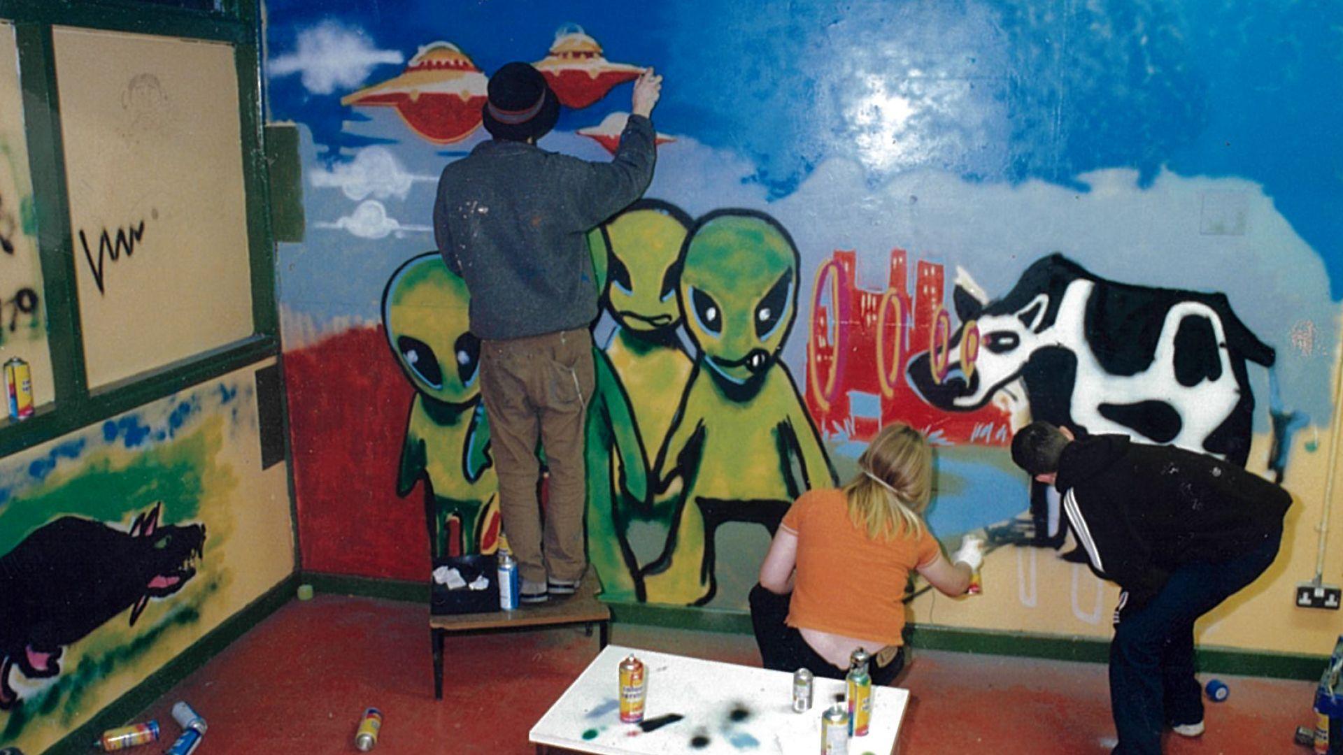 Banksy spray paints a spaceship on a wall, while two children add details to a cow with a series of circles coming from its eyes towards a group of green aliens.