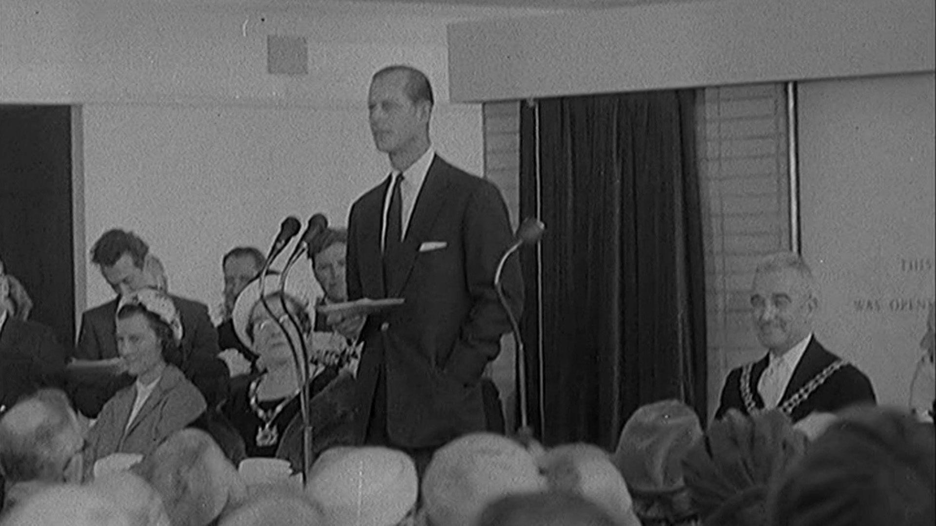 Black and white footage of Prince Philip opening the terminal. He stands in front of a crowd reading a speech. 