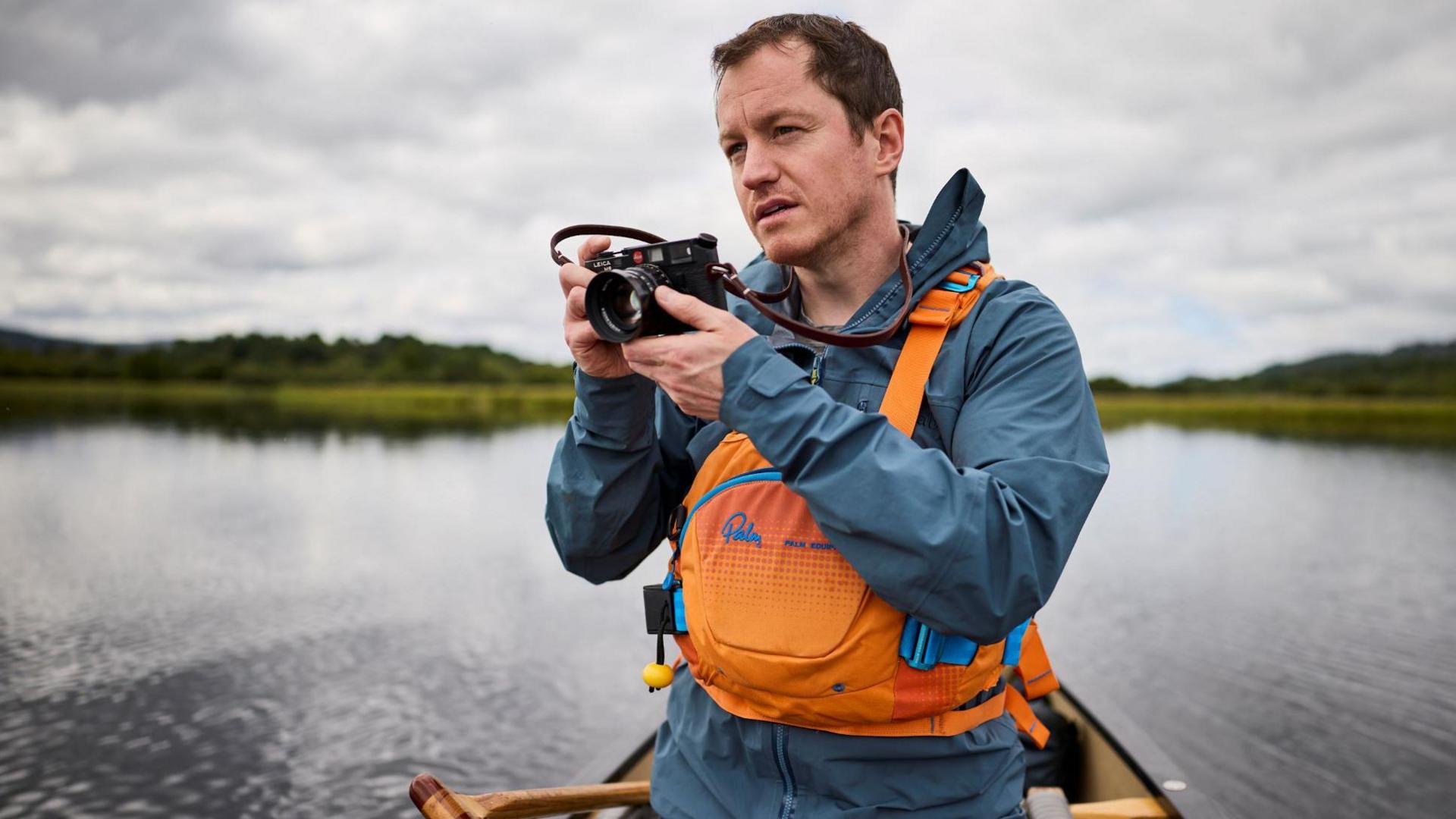 Ed is in a canoe on a loch. He is wearing an orange lifejacket over a blue outdoor jacket. Ed has brown hair and his holding a camera. He is looking off to the distance, framing a shot.