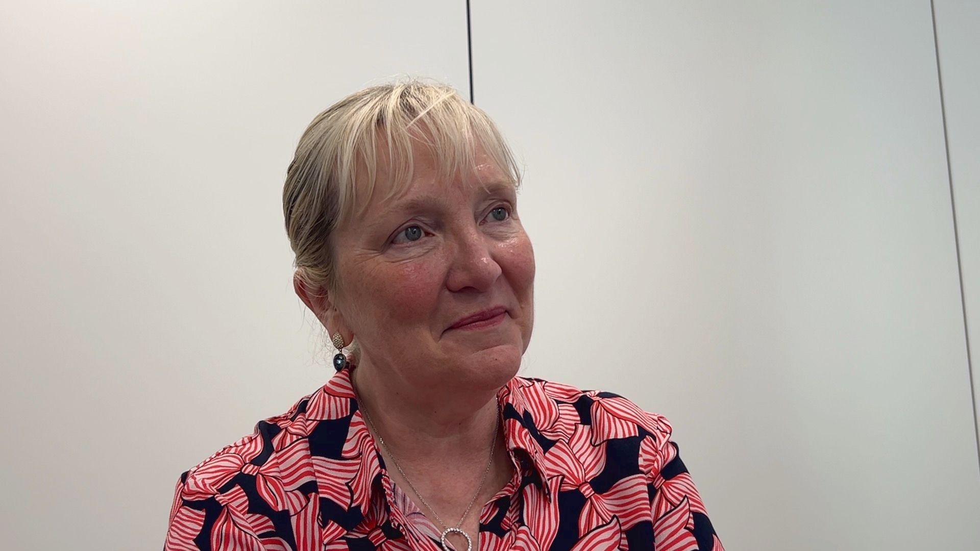Tricia Warwick is wearing a pink and black shirt. She has blonde hair and is wearing a necklace.