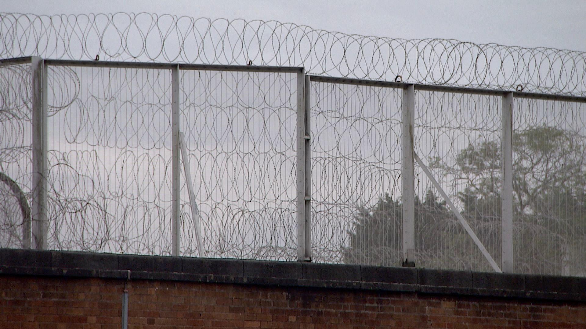 A barbed wire fence at Swinfen Hall prison. It is set against a backdrop of a grey sky. We can also see part of a high wall. 