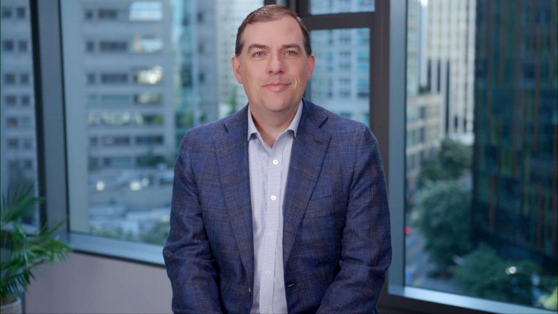 A medium close up of Matt Garman, chief executive Amazon Web Services, sat facing the camera in front of a window of an office building with the city outside visible. He is smiling slightly and wearing a blue suit and a shirt without a tie.